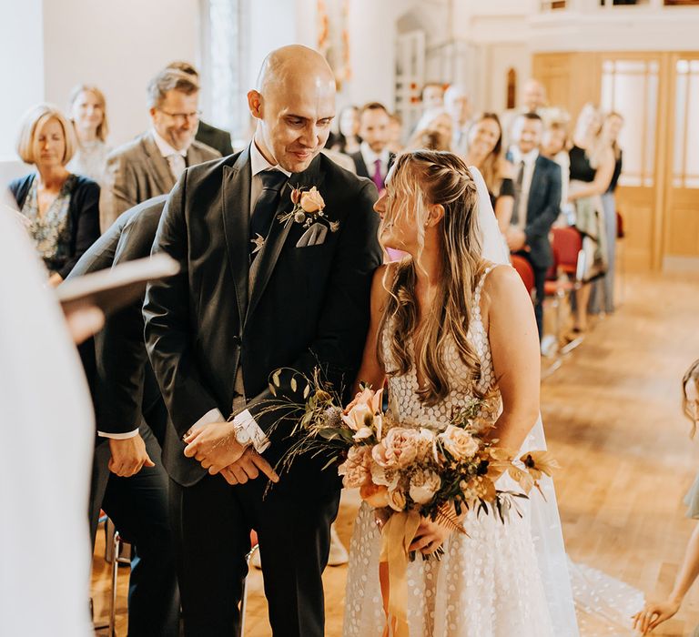 The groom in a dark suit stands at the altar with the bride holding a rustic autumnal bouquet with roses tied with orange ribbon smiling at each other 