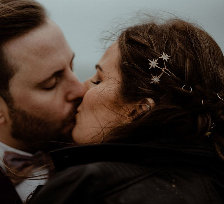 Groom in burgundy suit kisses the bride wearing a black leather jacket with gold star hair accessories 