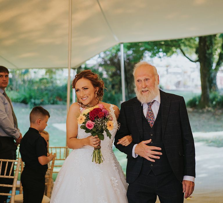 Bride walking down the aisle at a stretch tent wedding ceremony with colourful wedding bouquet