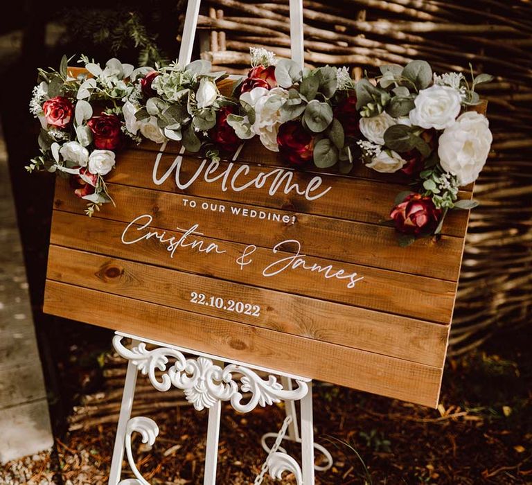 Wooden board welcome wedding sign with white cursive calligraphy on white stand with white and red garden rose, peony, eucalyptus and dried foliage flower arrangements on top 