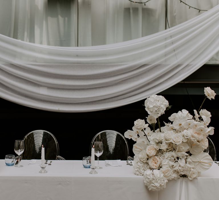 Sweetheart table for the bride and groom at their wedding breakfast decorated with white table cloth, white flowers and white drapery 