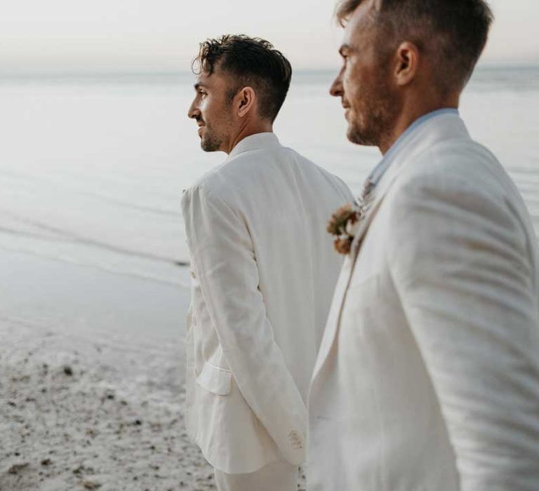 Grooms in complimenting double breasted white wedding suits holding hands doing couples photoshoot on the beach in Holbox 