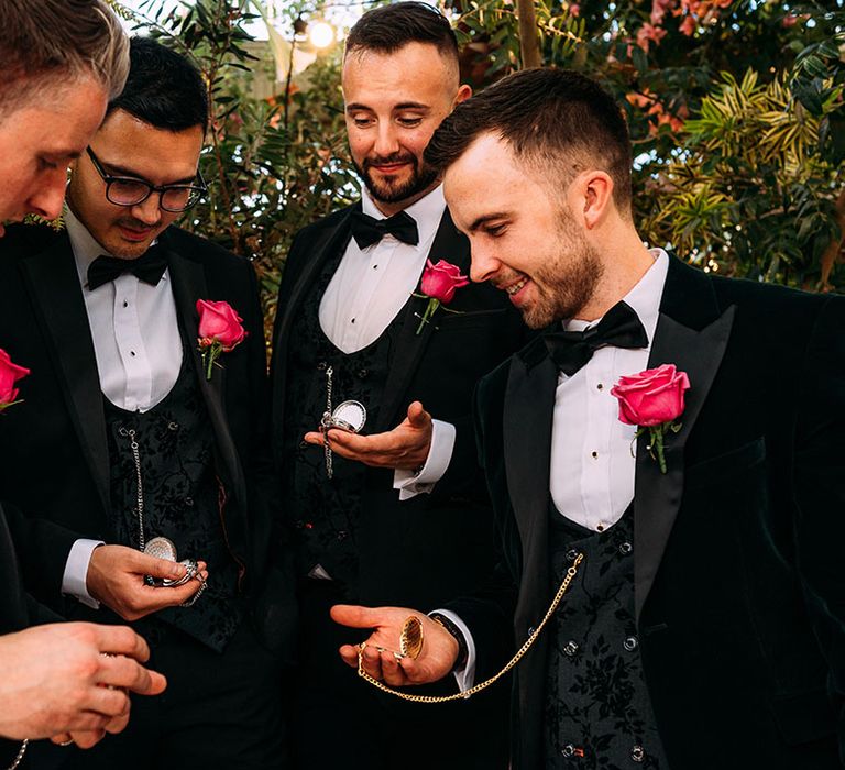 Grooms wearing matching black tuxedos with pink rose buttonholes holding pocket watches for the wedding day 