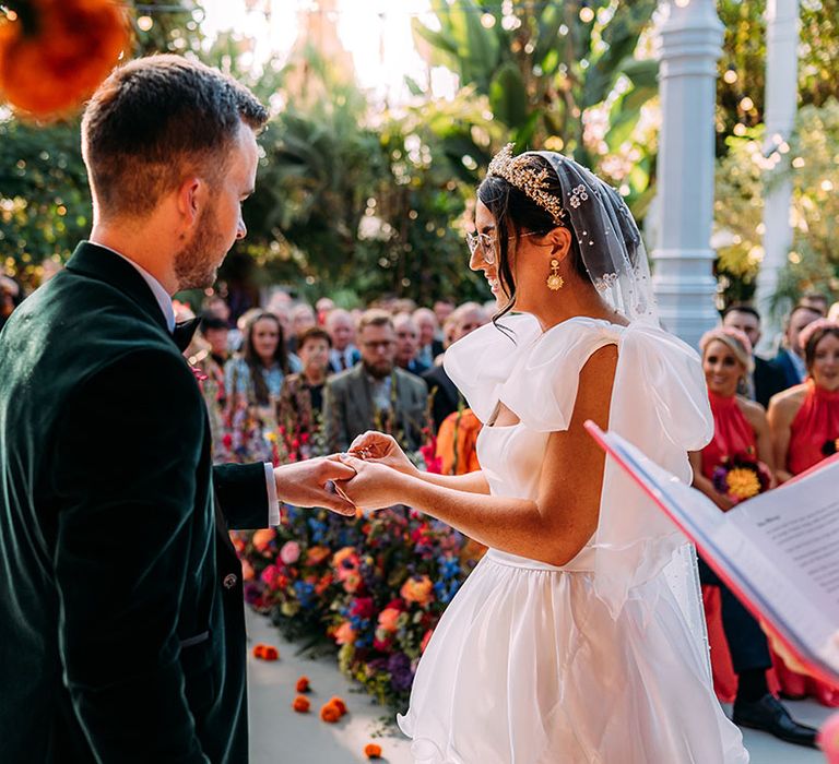 Bride wearing statement bow strap wedding dress with gold crown and beaded veil placing the wedding ring on the groom's finger 