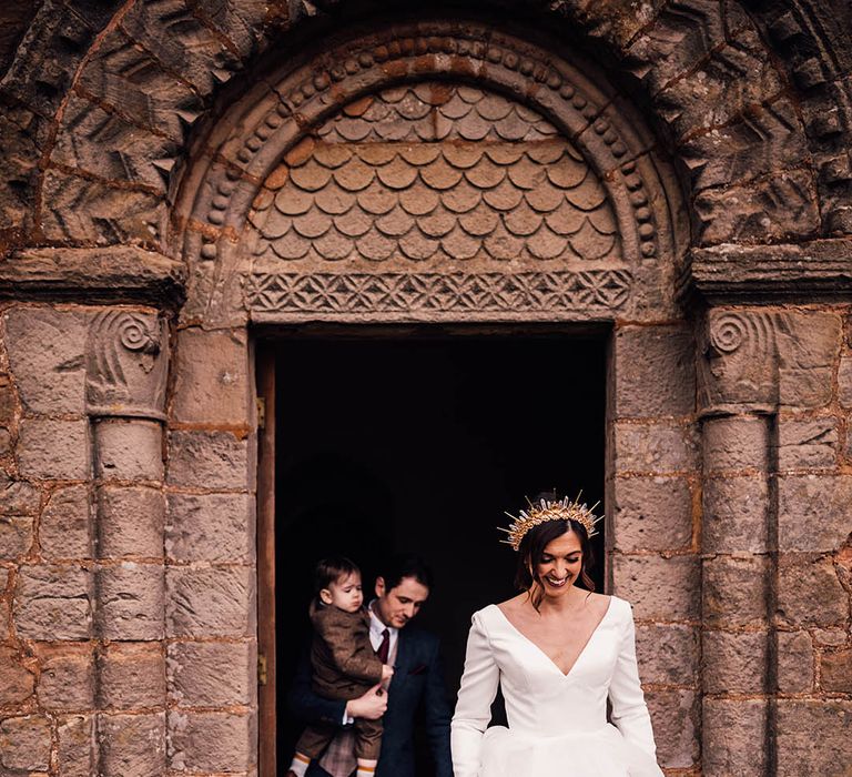 Bride in a ruffle gown and crown walking out of their wedding ceremony with the groom carrying their son 