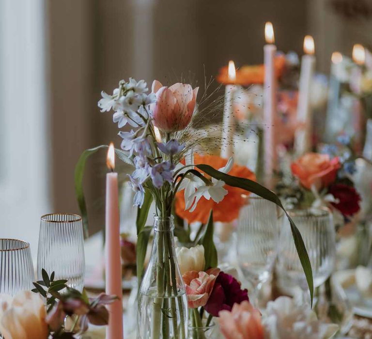 Spring themed wedding tablescape with orange poppies, pink and red roses, jasmine and bluebells on white wedding tablecloth and off-white satin table runner with pastel tapered candles, gold-rimmed wine glasses and classic crockery 