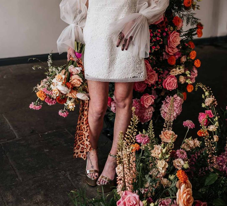 Bride wearing 70s-inspired boatneck short lace wedding dress with detachable tulle puff sleeves and platform gold wedding heels standing by circular floral arch with vibrant flowers and foliage attached