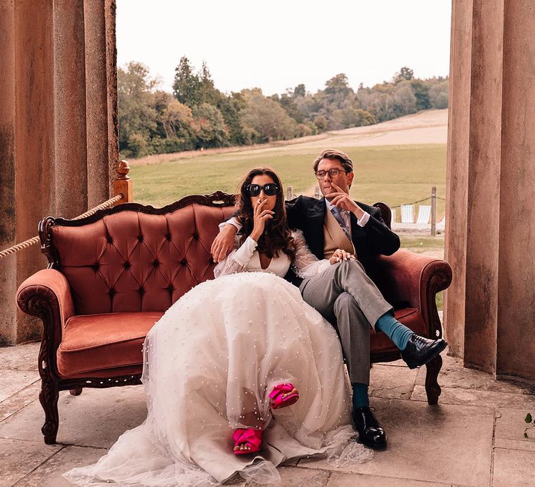 The bride and groom sit leisurely on the pink sofa enjoying a cigarette for their Wes Anderson and Gatsby inspired wedding 