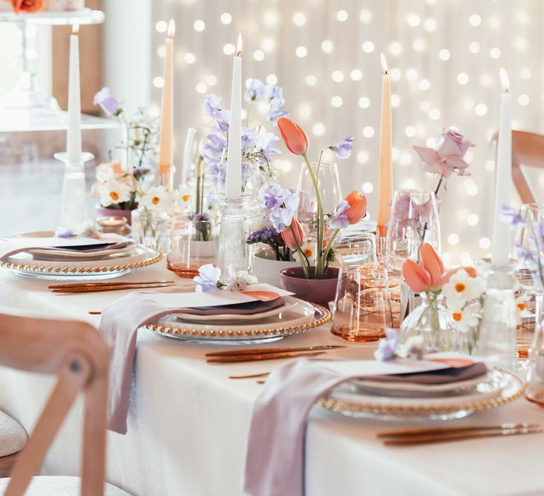 Peach and lilac wedding tablescape with spring flower centrepieces, lilac napkins, tinted glassware and gold cutlery and crockery on white tablecloth with wedding fairy lights wall at Rackleys Barn