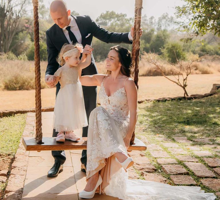 Wedding family photoshoot - groom in classic black grooms suit with white pocket square helping child in off-white dress and sequin headband with pink carnation standing next to bride in lace sleeveless wedding dress with puddle train