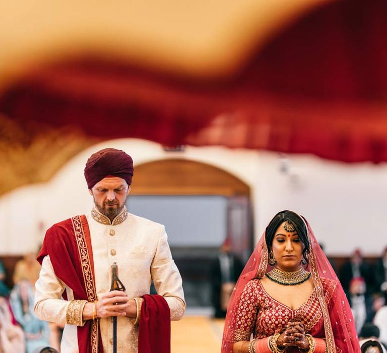 Groom in white and gold sherwani doing Sikh wedding ceremony with bride in red and gold lehenga and gold and forest green bridal jewellery  