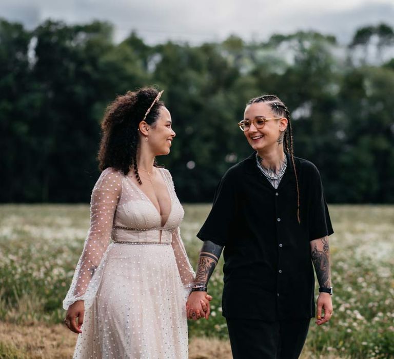 Bride in long sleeve v-neck sheer polka dot overlay sparkly wedding dress and gold celestial bridal headband walking hand in hand with bride in silver chains, glasses, black shirt and black trousers at Viking themed wedding