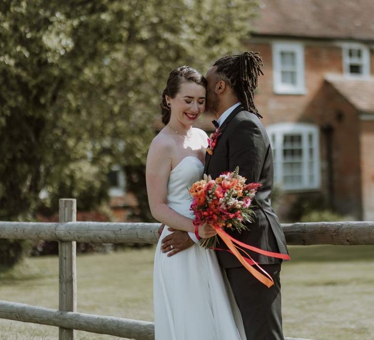 Bride in strapless sweetheart neckline wedding dress holding pink and orange wedding bouquet with groom in classic black tuxedo with bowtie and pink and orange wedding boutonniere at The Old Rectory Berkshire