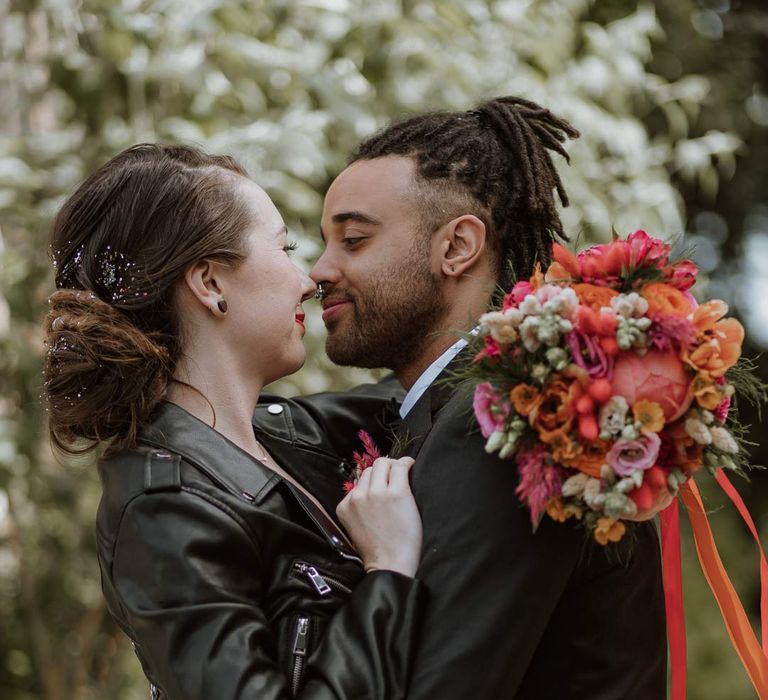 Bride in strapless sweetheart neckline wedding dress and personalised bridal leather jacket embracing groom in classic black tuxedo 