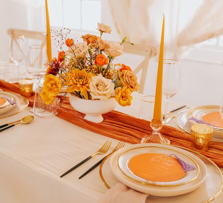 Orange rose bowl flower arrangement with orange taper candles, table runner and circular wedding menu 