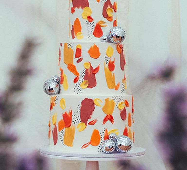 White iced three tier wedding cake with red, yellow and orange painted style icing with disco balls 