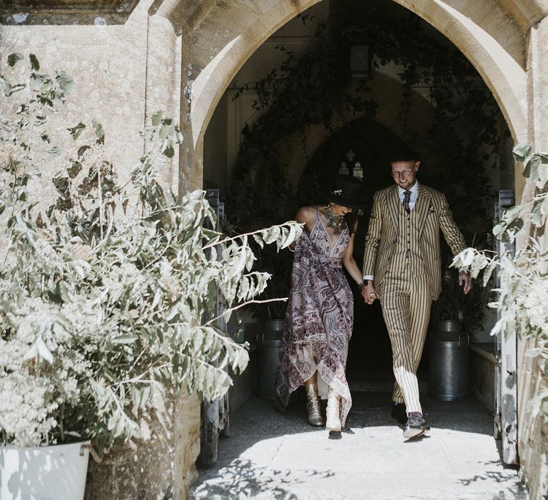 bride in pink bohemian wedding dress, silver boots and wedding hate holds hands with the groom in pinstripe groom suit exiting church wedding ceremony