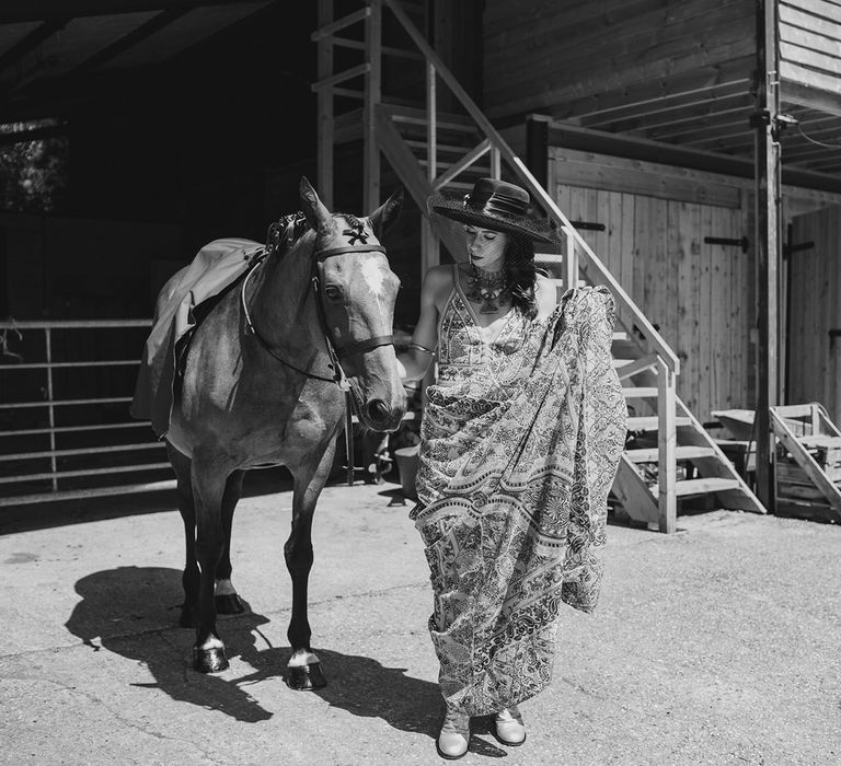 Bride in boho wedding dress walks with horse outside stables