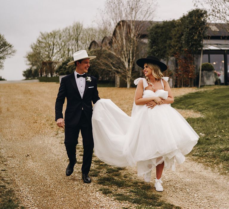 Bride wearing white wedding trainers in princess wedding dress with black cowboy hat walking with groom in tux with white cowboy hat