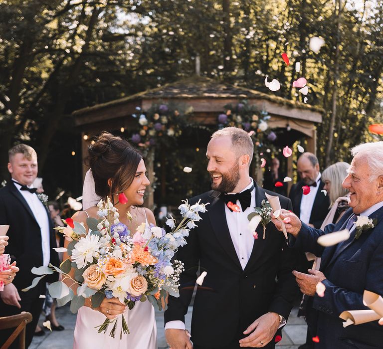 Bride in satin slip wedding dress walking with groom out to colourful confetti exit 