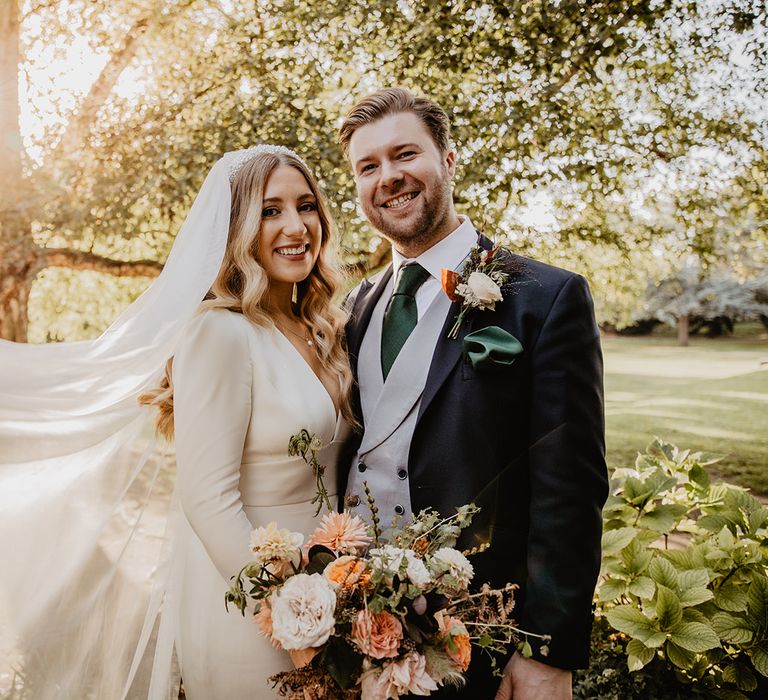 Bride in long sleeve wedding dress and veil smiling with groom in blue three piece suit with grey waistcoat and dark green satin tie 