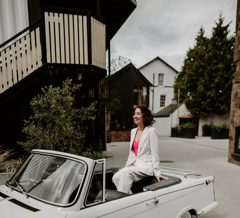 Bride in white blazer riding in vintage convertible wedding car 