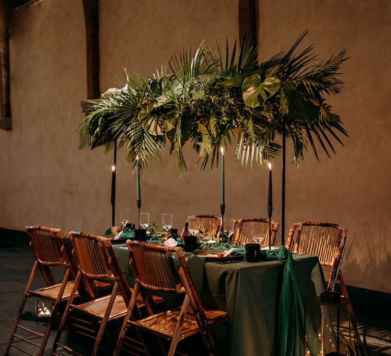 Dark green wedding tablecloth with candle lanterns, wooden chairs, and palm leaves foliage installation 