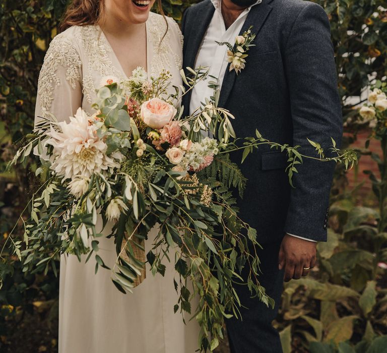 Boho bride in ivory gown and oversized bouquet and groom in lounge suit 