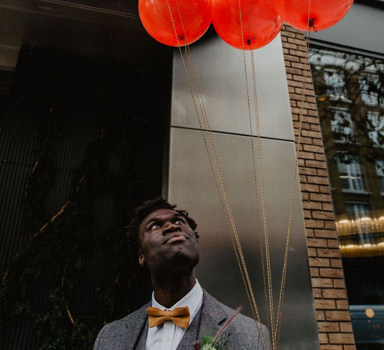 Black Groom wearing mustard bow tie holds bunch of red I love you balloons