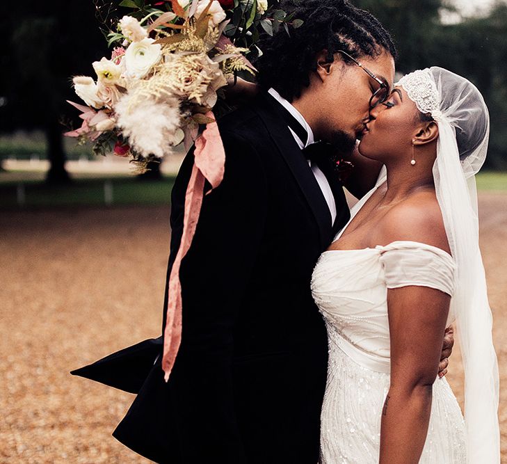 A bride and groom kiss. The bride wears aJuliet Cap veil and has cropped hair underneath.