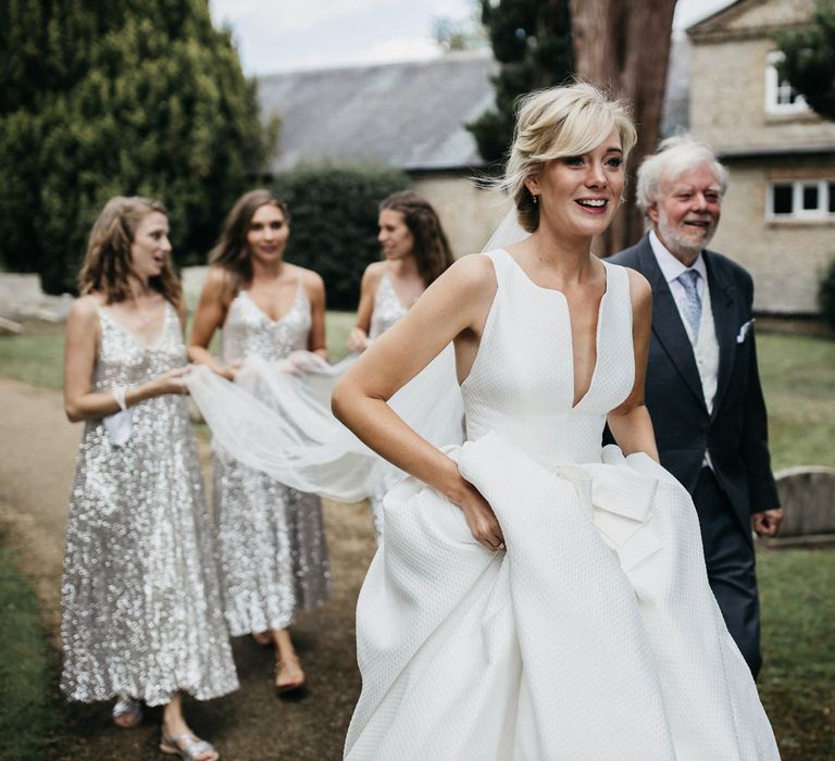 Bride in a Jesus Peiro wedding dress with plunging neckline walking through the church courtyard with her father and bridesmaids 