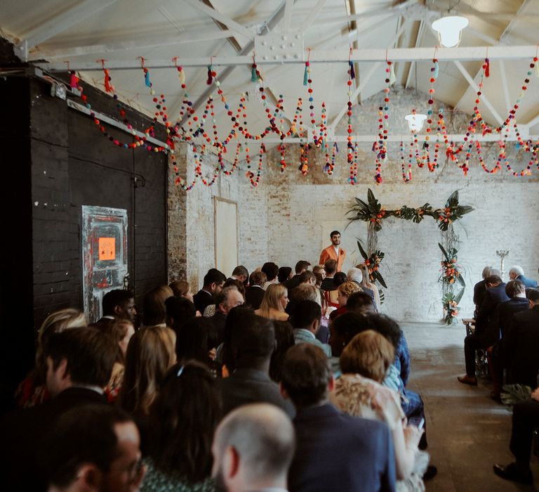 Turkish groom in a double breasted orange suit standing at the altar of industrial wedding venue decorated with colourful pompom ceiling decor and tropical flower altar