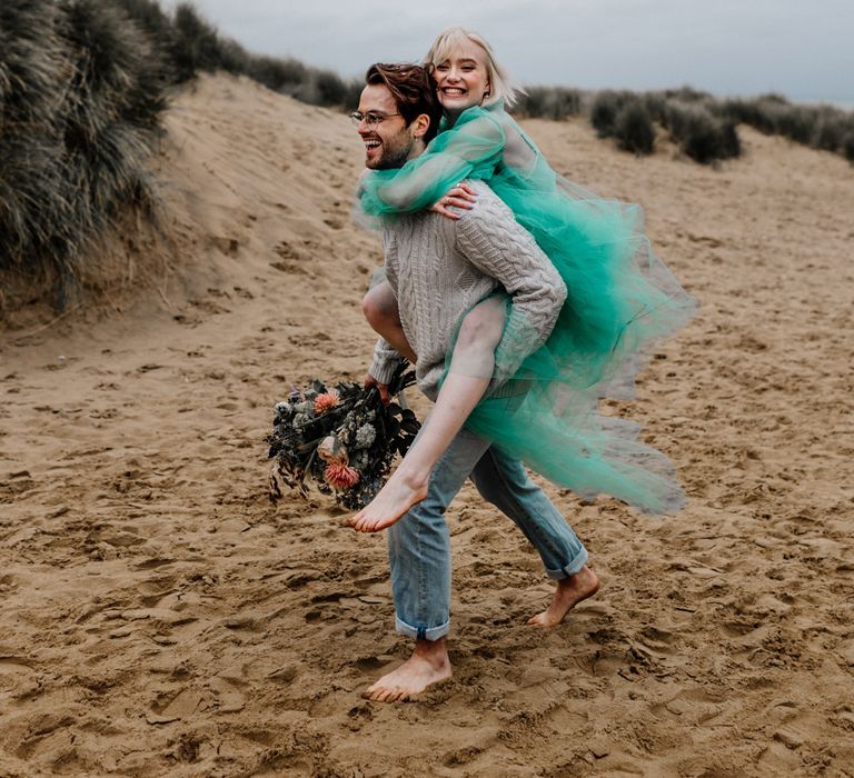 Bride in a sheer green wedding dress having a piggy back from her groom in jumper and jeans 