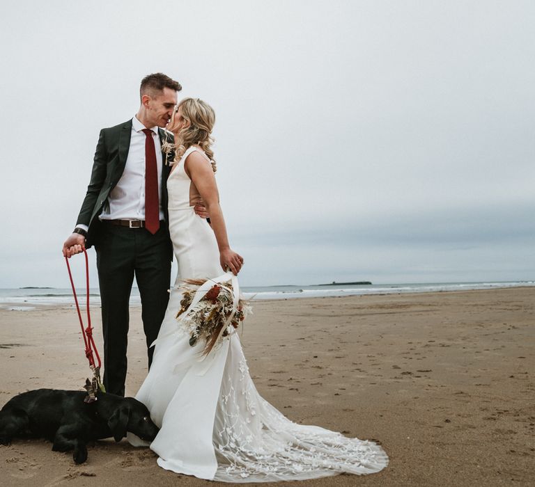 Bride & groom stand together on the beach whilst their dog lies at their feet