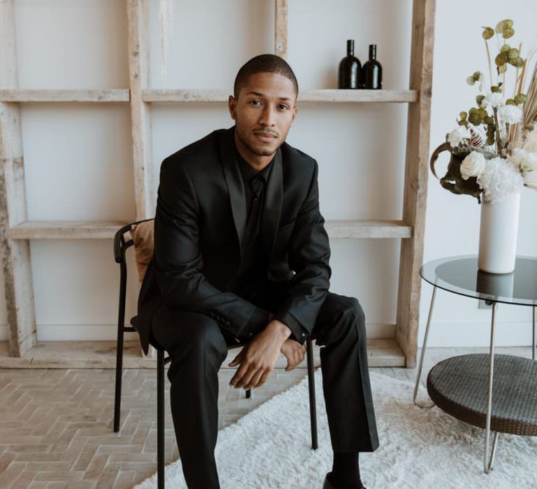 Black groom sitting on chair in all black suit and black shirt