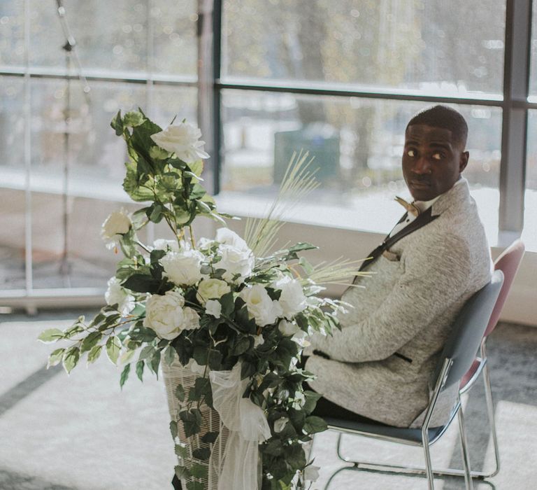 Groom in grey Moss Bros suit sits in blue chair next to white floral installation waiting for bride at Bridge Community Church