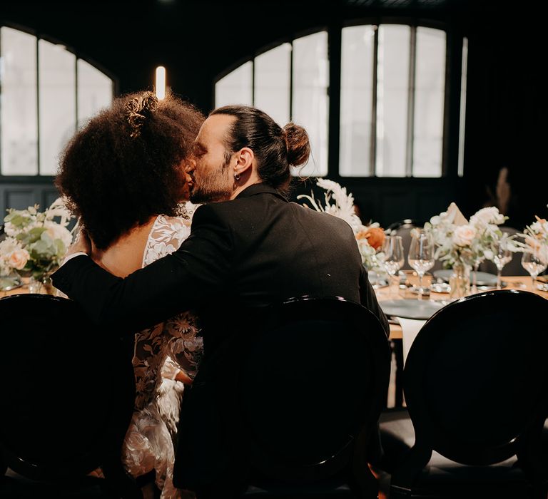 Bride & groom kiss during reception dinner in front of windows 