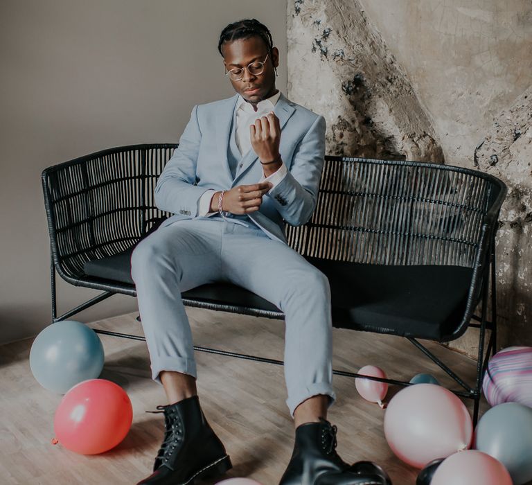 Stylish groom in a baby blue wedding suit and boots sitting on a black love seat surrounded by pastel balloons 