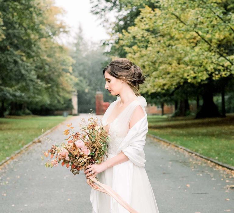 Bride in a tulle and lace wedding dress holding an orange flower autumn bouquet wrapped in a cream shawl 