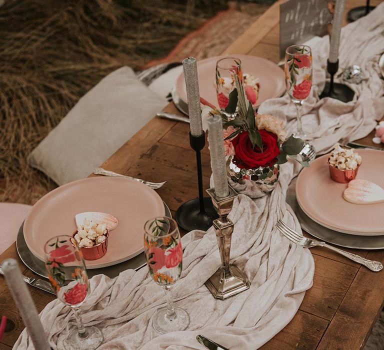 Tablescape at festival themed wedding with white velvet table runner on wooden table, tropical print champagne flutes, candle and pastel pink plates