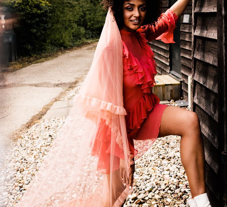 Bride in a short red wedding dress and long pink veil with ruffles standing next to her barn wedding venue 