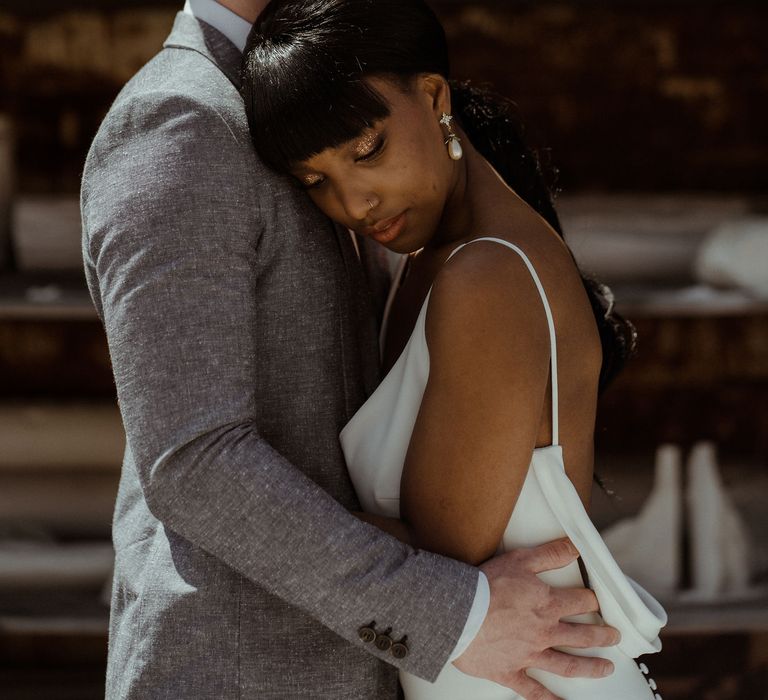 A couple hug. He wears a grey suit and she wears a low back white wedding dress. 