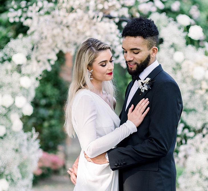 Groom in a tuxedo embracing his bride in a one shoulder long sleeve wedding dress with wrap skirt 