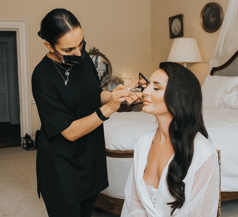 Bride with long dark curled hair in white satin bridal dressing gown has her make up done by woman in black t shirt and black face mask in bedroom at Came House Dorset