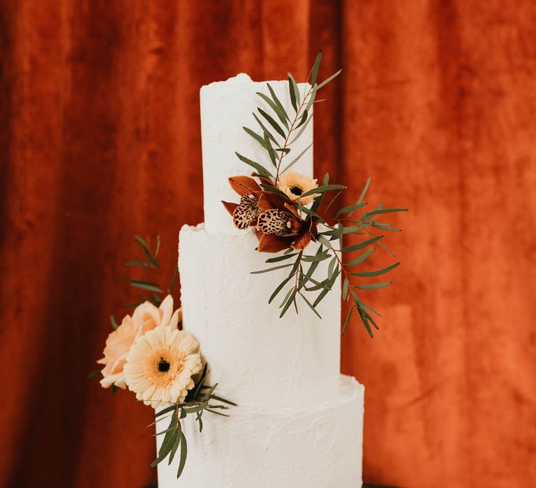 Three tier plain white wedding cake by Where The Ribbon Ends with peach and coral flower decoration, in front of coral red backdrop