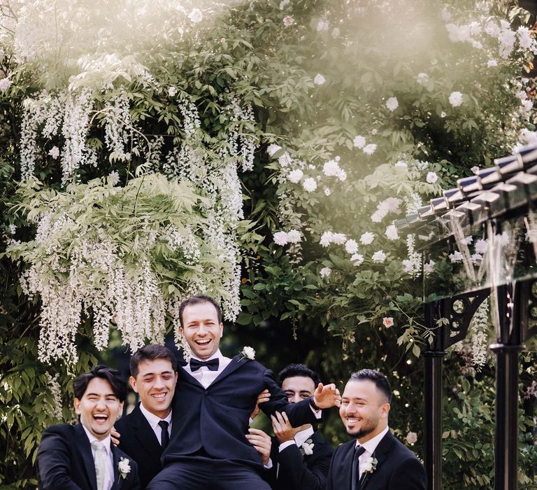 Groom held up by his groomsmen in matching black tie suits under hanging wisteria