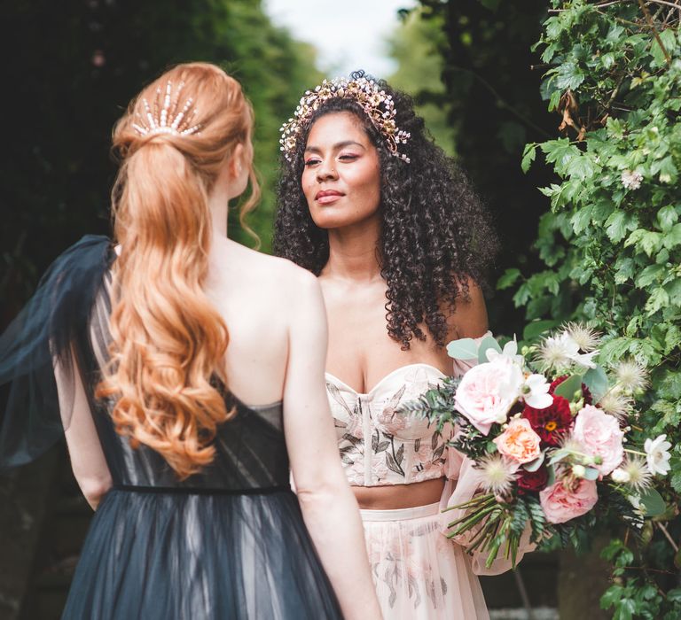 Black bride with long naturally curly hair wearing a bridal headdress in an embroidered bodice and tulle skirt looking at her bride-to-be