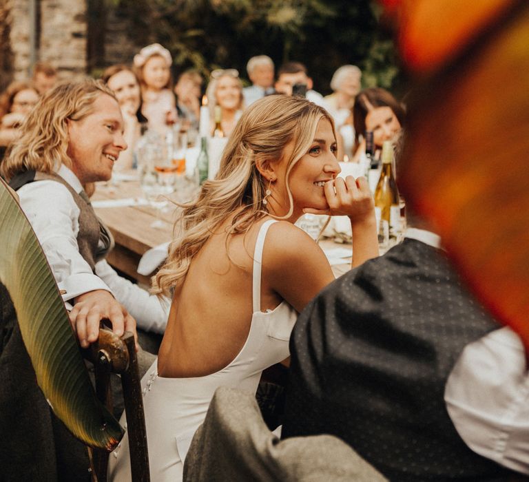 Smiling bride sits next to smiling groom at wooden wedding breakfast table in glasshouse at Anran Devon as they watch speeches 