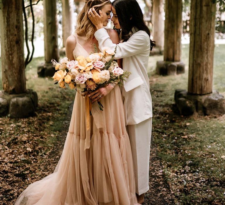 Bride in a white suit embracing her brides face in a blush pink tulle wedding dress as she holds a peeled back rose wedding bouquet 