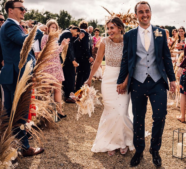 Bride in white Rime Arodaky wedding dress with lace top holding dried bridal bouquet smiles as she walks down the aisle holding hands with groom in blue three piece suit and dried floral buttonhole as guests throw confetti after outdoor wedding ceremony in Dorset - Carrie Lavers Photography